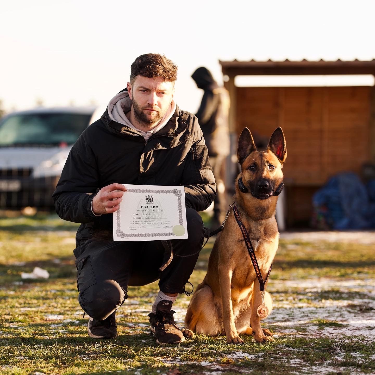 Jason Cooper kneeling beside his Belgian Malinois, holding a PSA certificate at a trial field