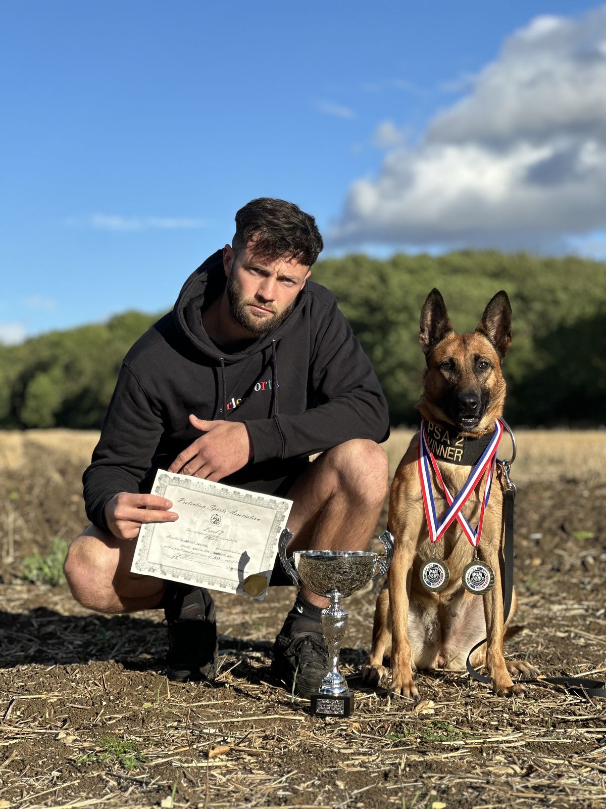 Jason crouching beside his Malinois, who wears two medals, with a trophy and PSA certificate