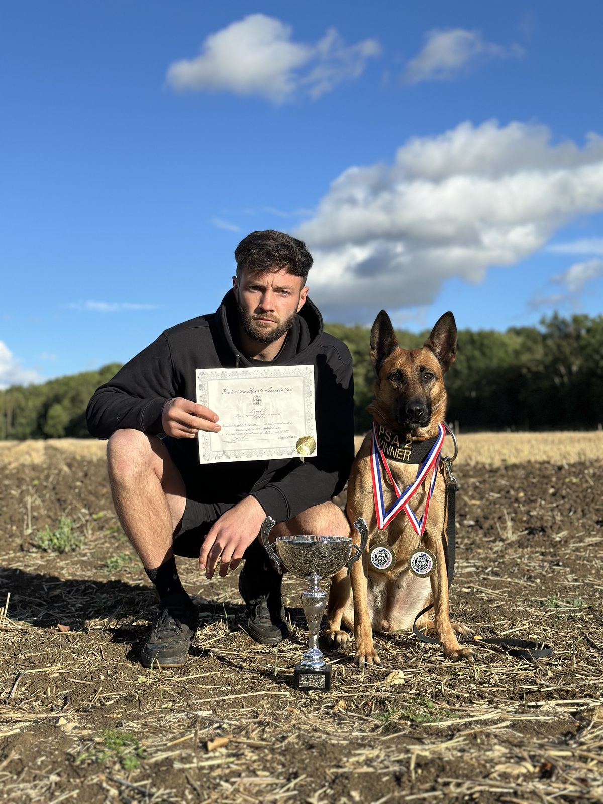 Jason crouching with his Malinois, a PSA certificate, trophy and medals in a stubble field
