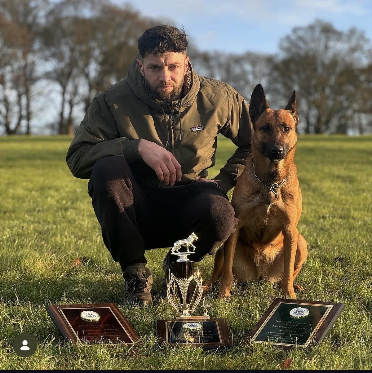 Jason and his Malinois on grass behind a trophy and two presentation plaques