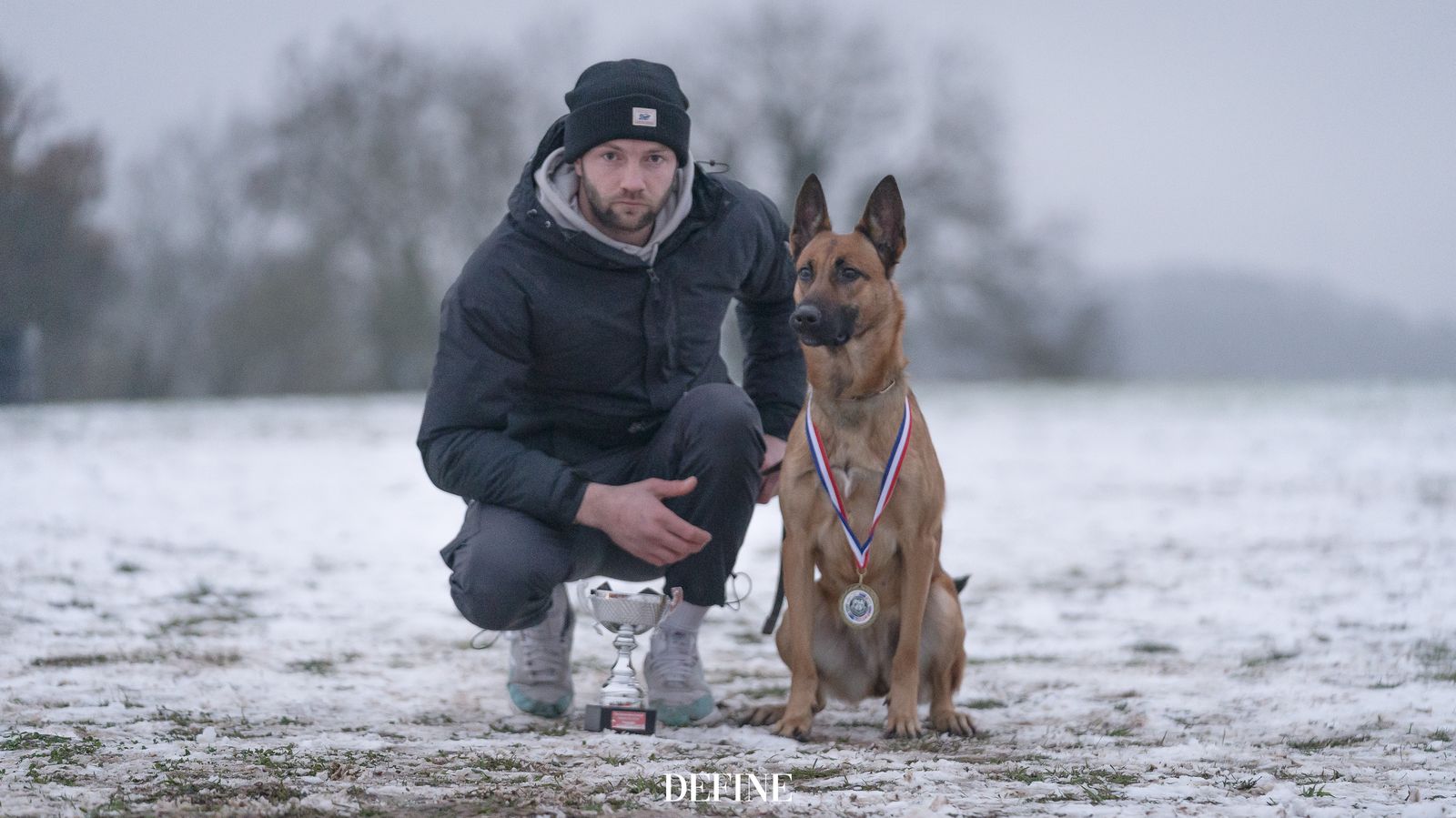 Jason kneeling in a snowy field next to his Malinois, who wears a medal beside a trophy